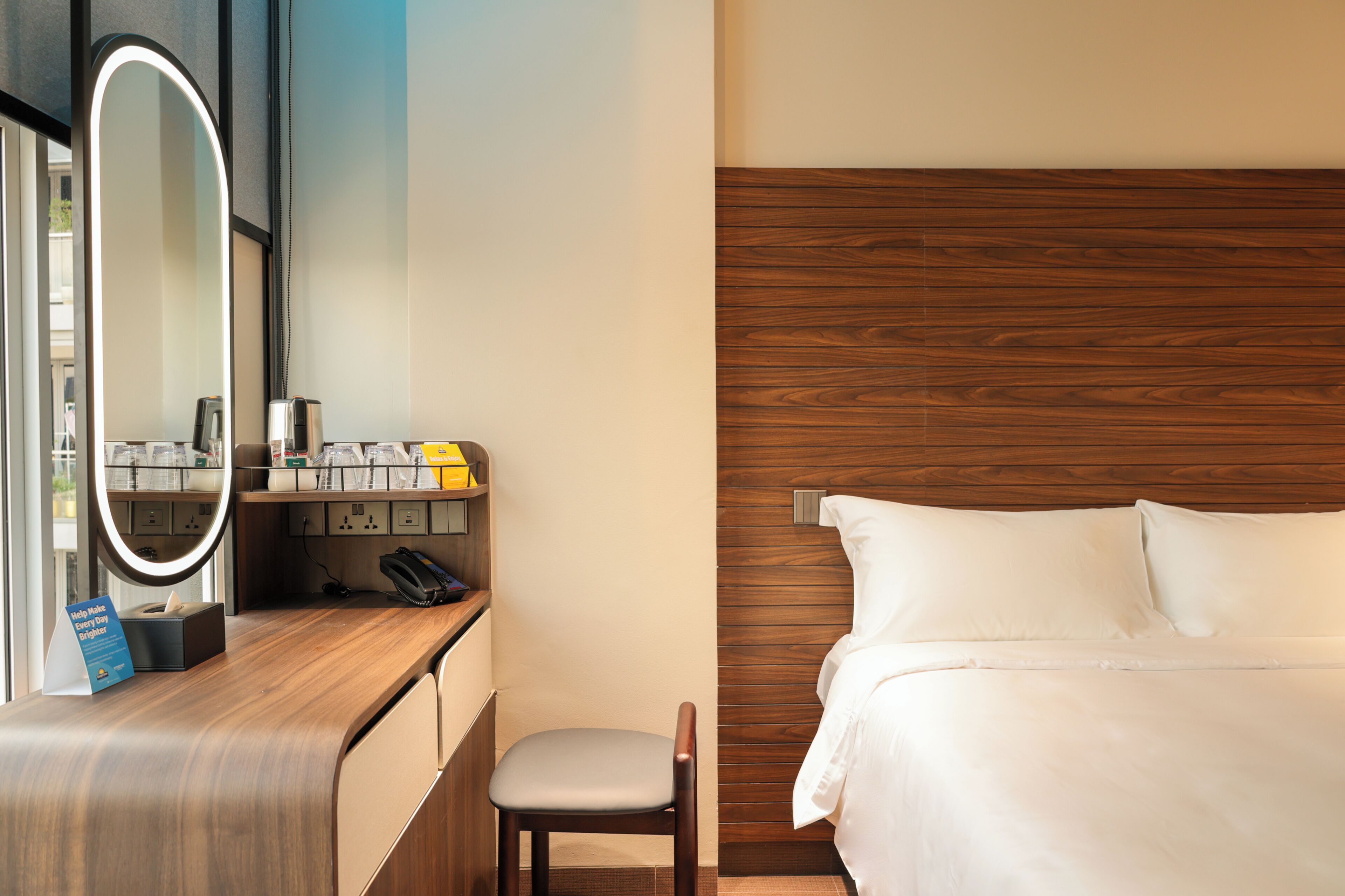 A hotel room with a wooden vanity, a mirror, a bedside table with toiletries, a chair, and a bed with white linens.