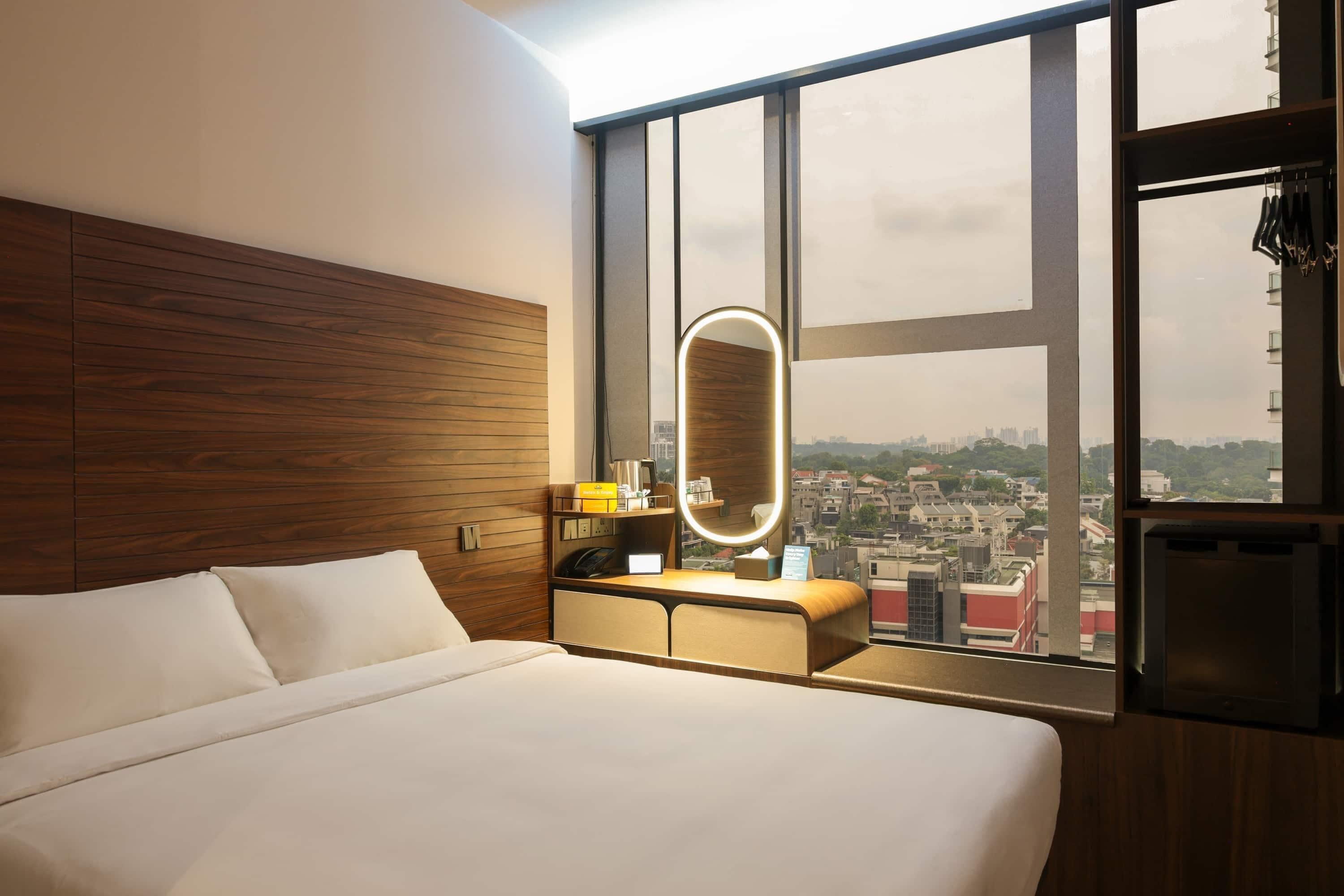 A hotel room with a large bed, a bedside table with a mirror, a dresser, and a view of a cityscape through large windows.
