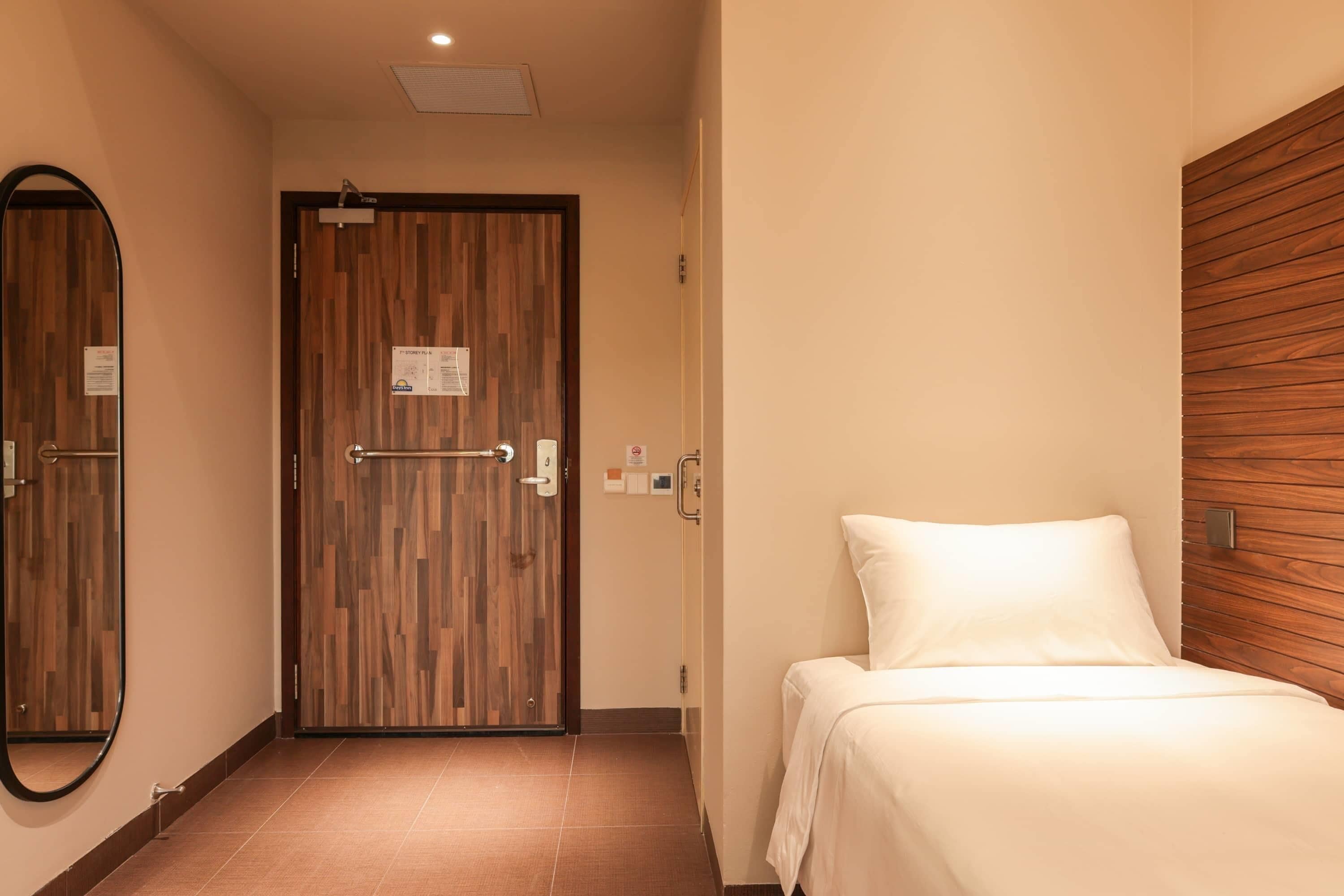 A hotel room with a wooden door, a bed with white bedding, a mirror, and a wall-mounted light fixture.