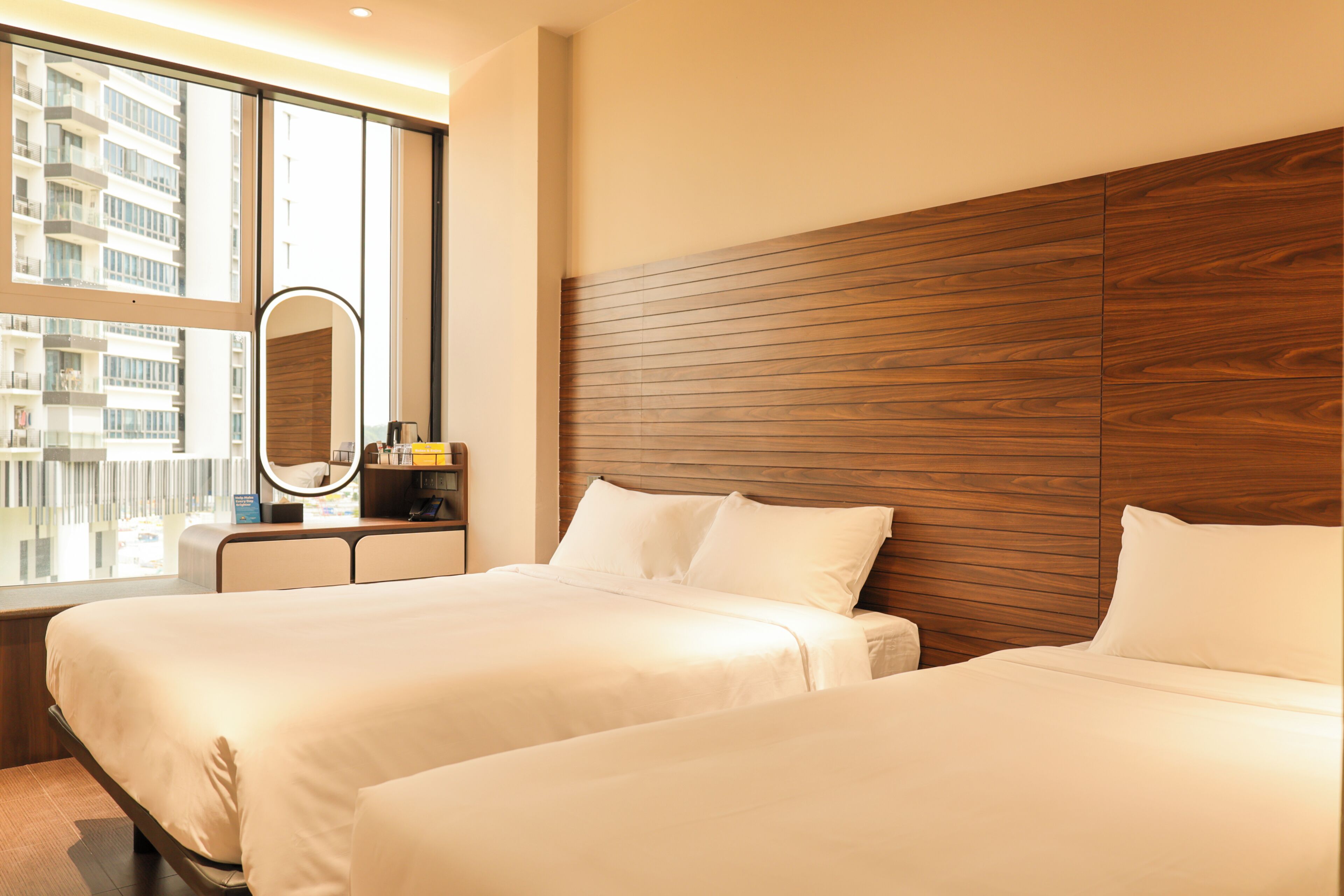 Une chambre d’hôtel avec deux lits, une tête de lit en bois, une grande fenêtre et un miroir rond.