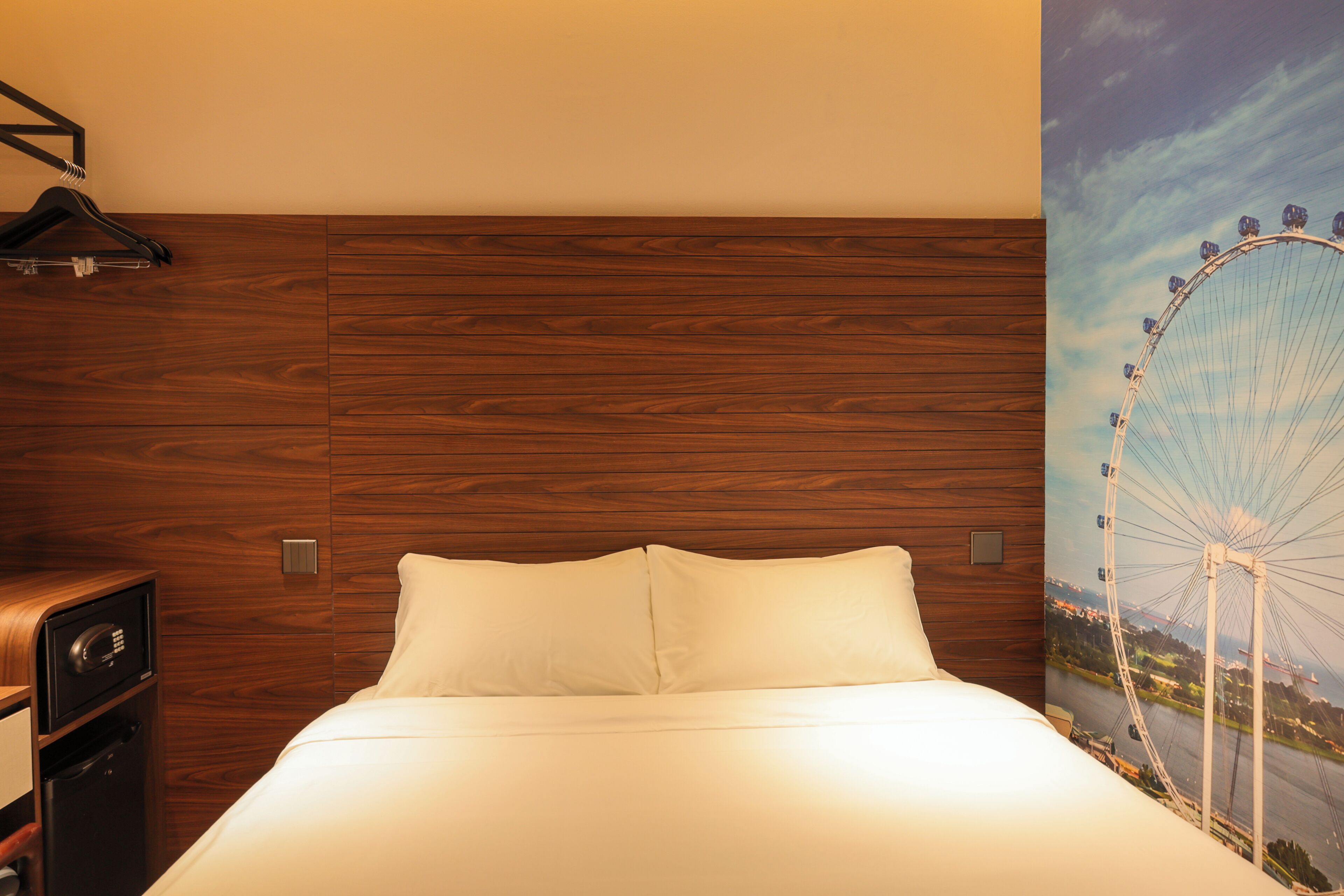 A hotel room with a wooden headboard, a bed with white linens, a wall-mounted safe, and a large window with a view of a Ferris wheel.