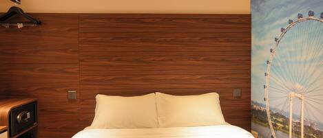Habitación de hotel con cabecera de madera, cama con ropa blanca, caja fuerte en la pared y un ventanal con vista a una noria.