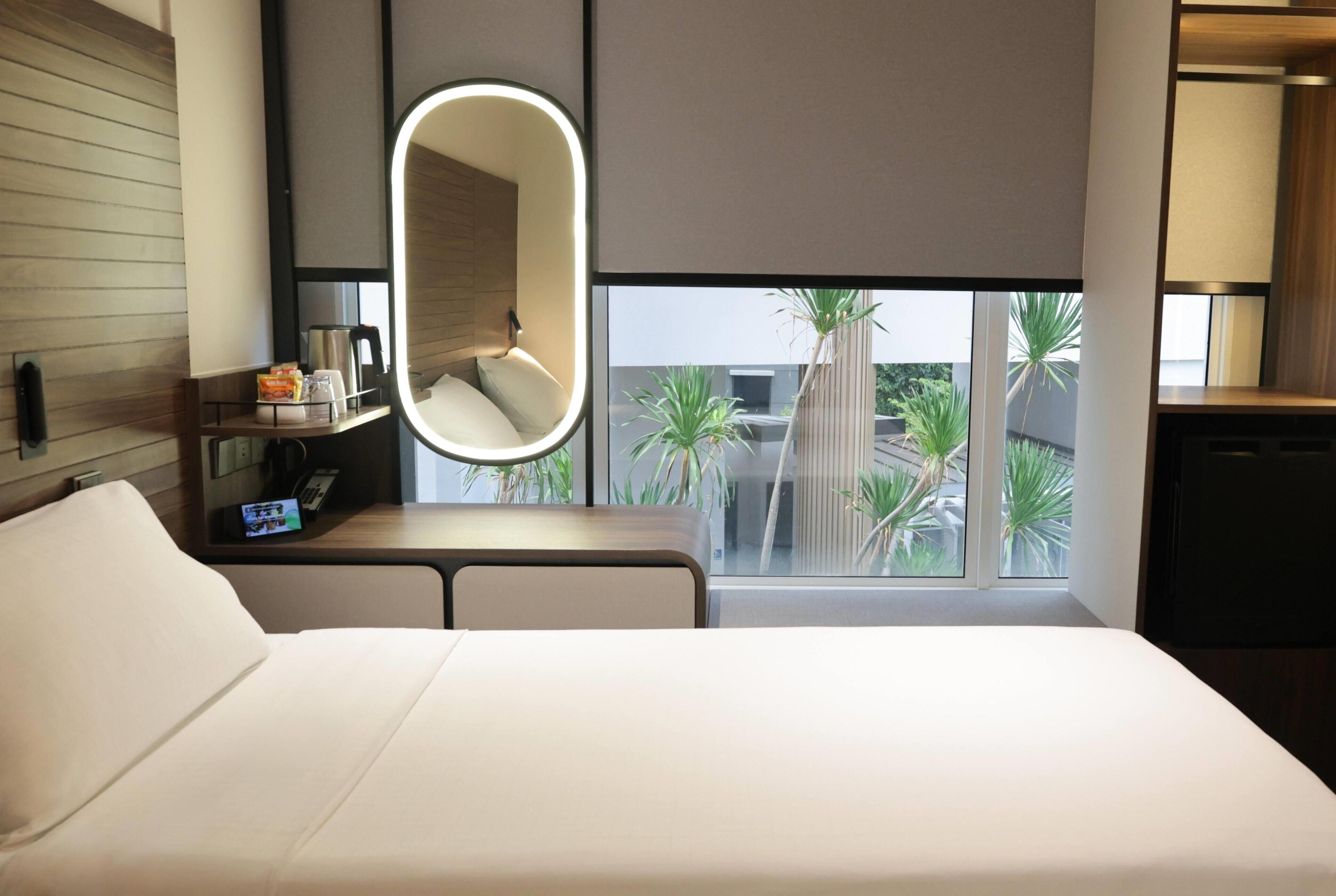 A hotel room with a bed, a bedside table with a coffee maker, a mirror, and a window with a view of palm trees.