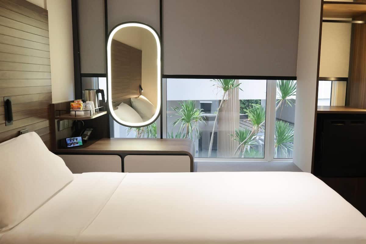 Habitación de hotel con cama, mesita de noche con cafetera, espejo y ventana con vistas a palmeras.