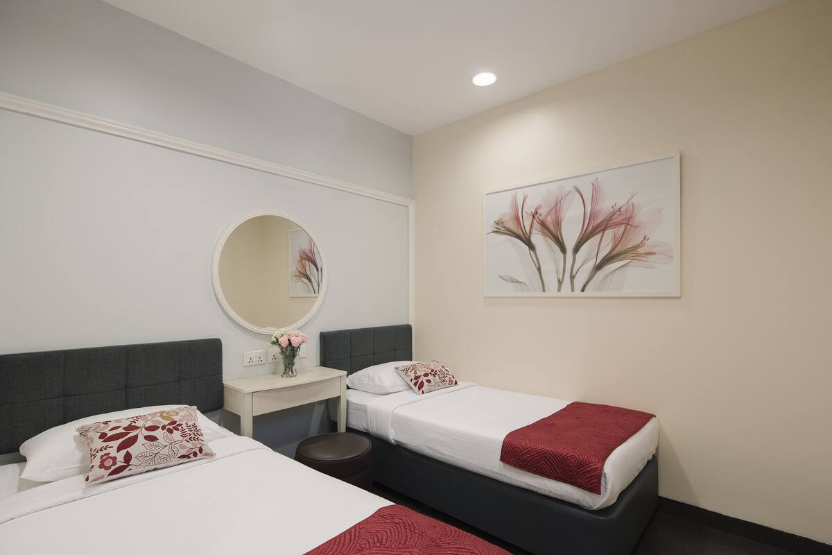A hotel room with two beds, a round mirror, a small table with a vase of flowers, and a painting on the wall.