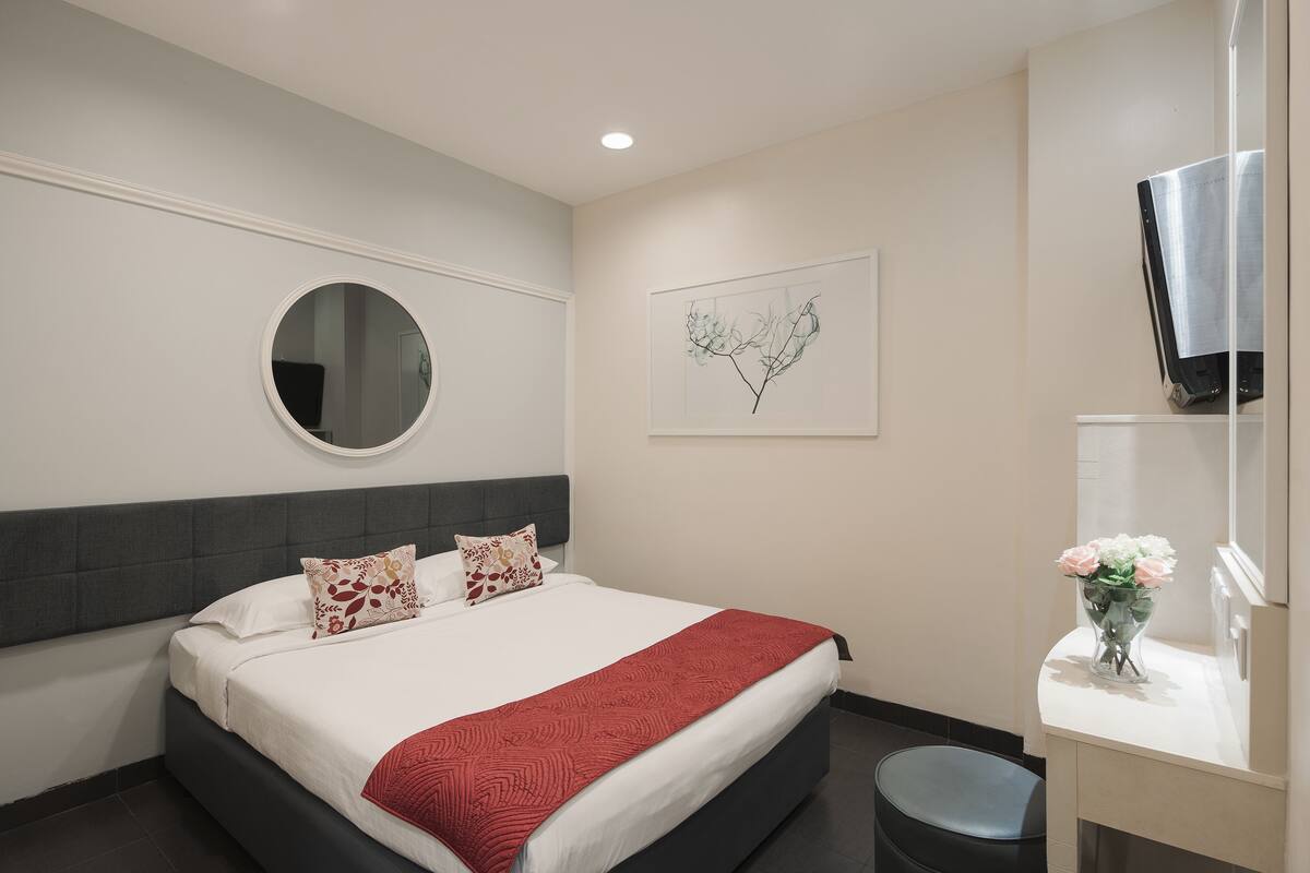 A hotel room with a bed, a round mirror, a wall art piece, and a small table with a vase of flowers.