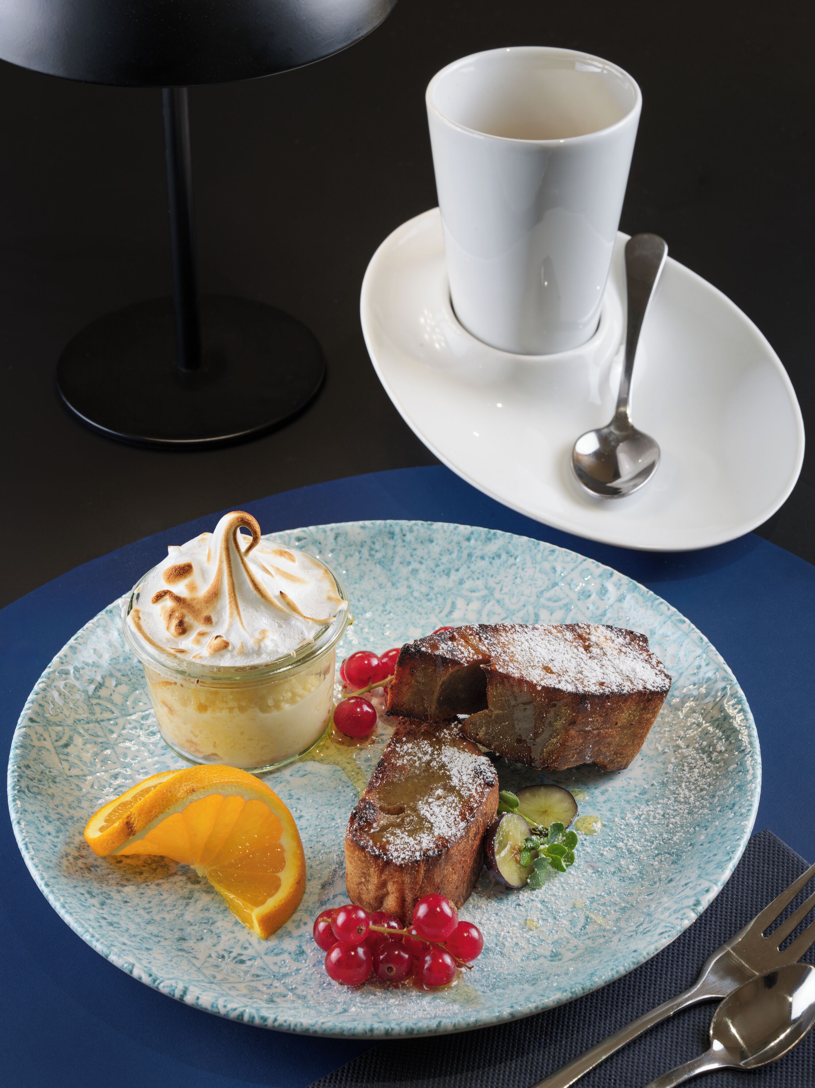 A dessert plate with a meringue, fruit, and powdered sugar, a white cup and saucer, and a spoon on a blue tablecloth.