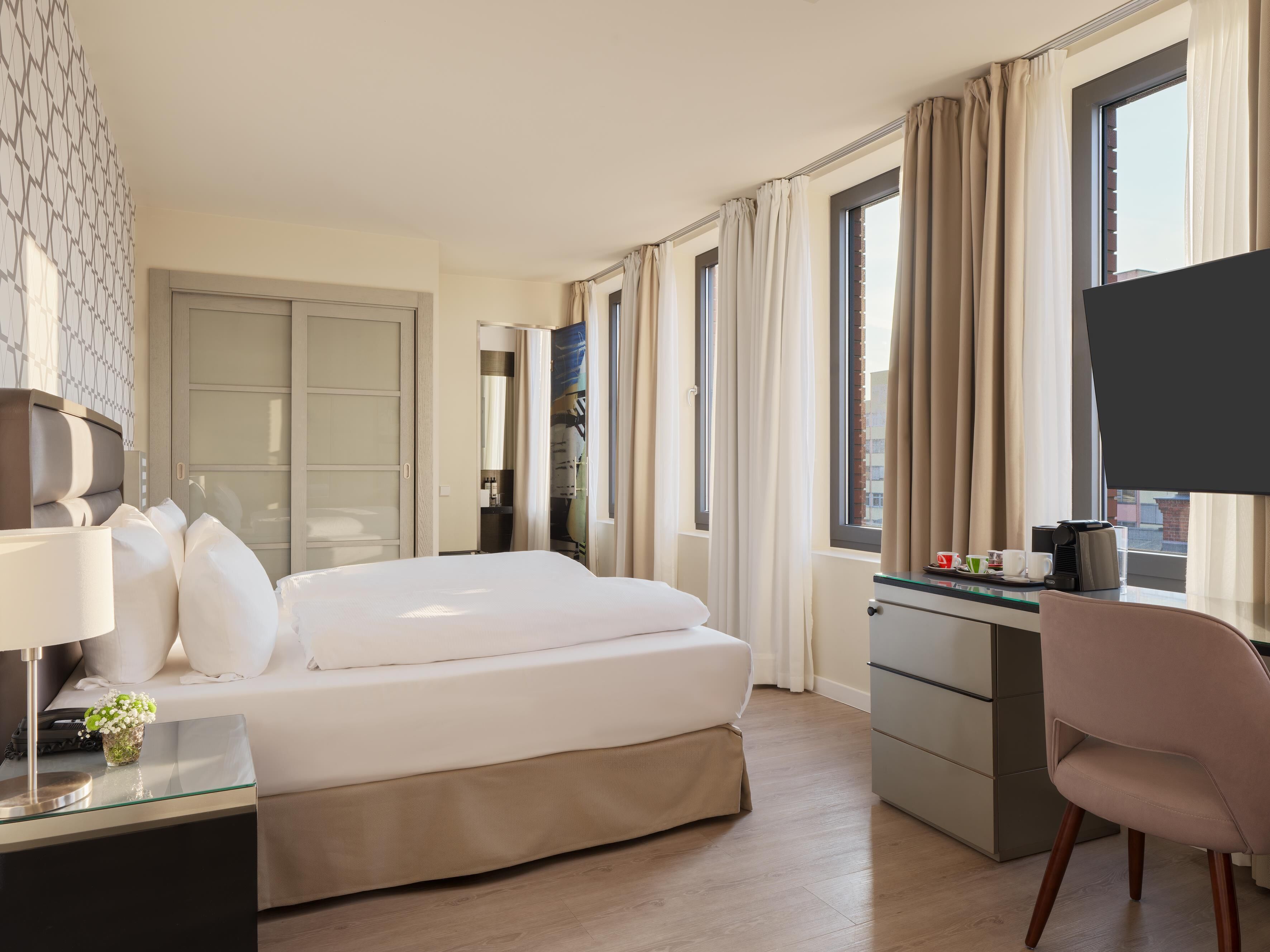 A hotel room with a bed, a desk, a chair, a TV, and a window with curtains.