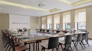 A conference room with long tables, chairs, a projector, and a screen displaying 'H1O Berlin Ku'damm'.