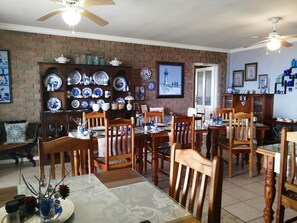 A dining area with a brick wall, wooden chairs, a glass table, and a variety of blue and white ceramic dishes displayed on shelves. - Amzee Bokmakierie Guest House (Mossel Bay)