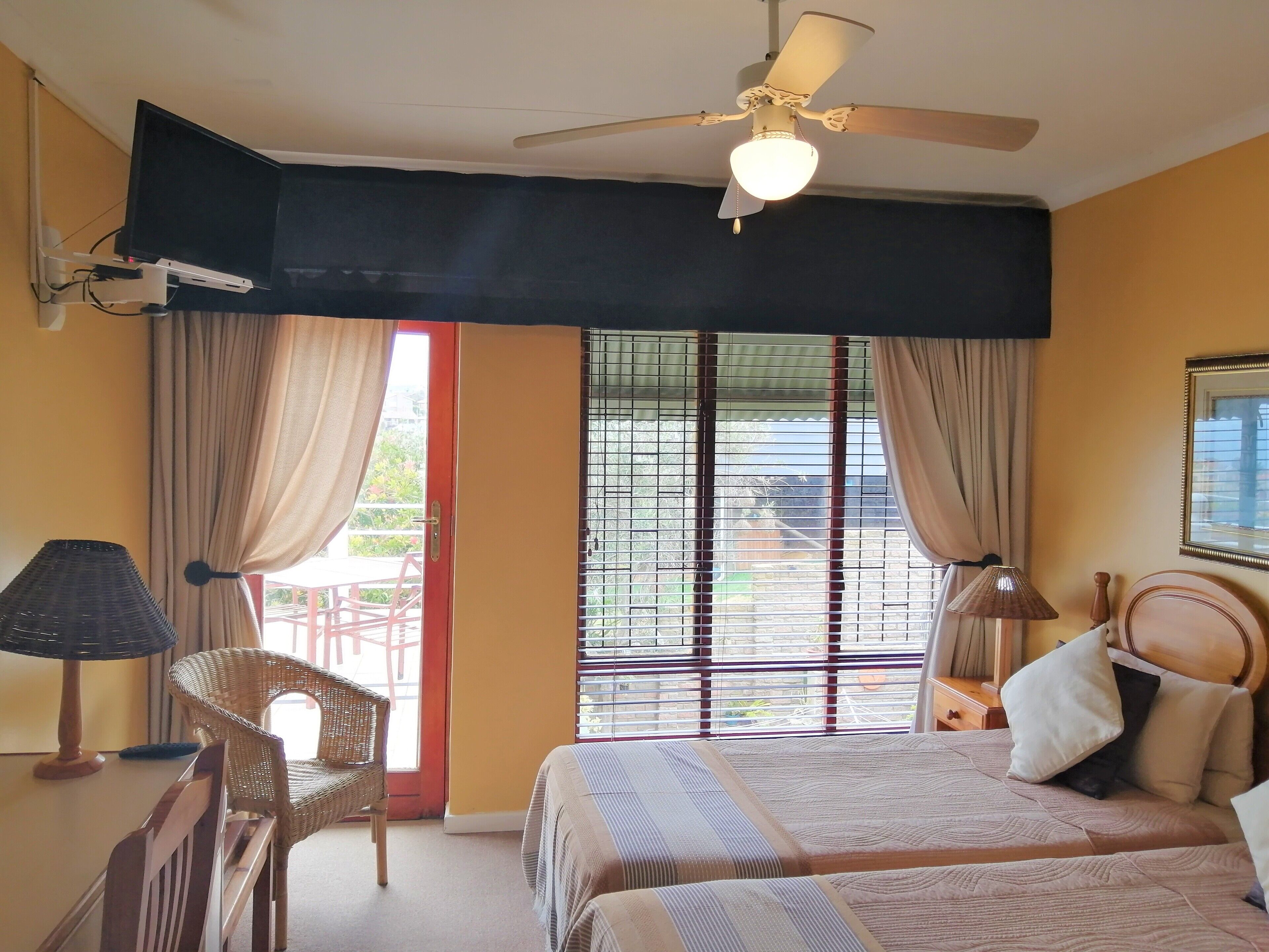 A hotel room with a bed, a chair, a desk, a ceiling fan, and a window with blinds.