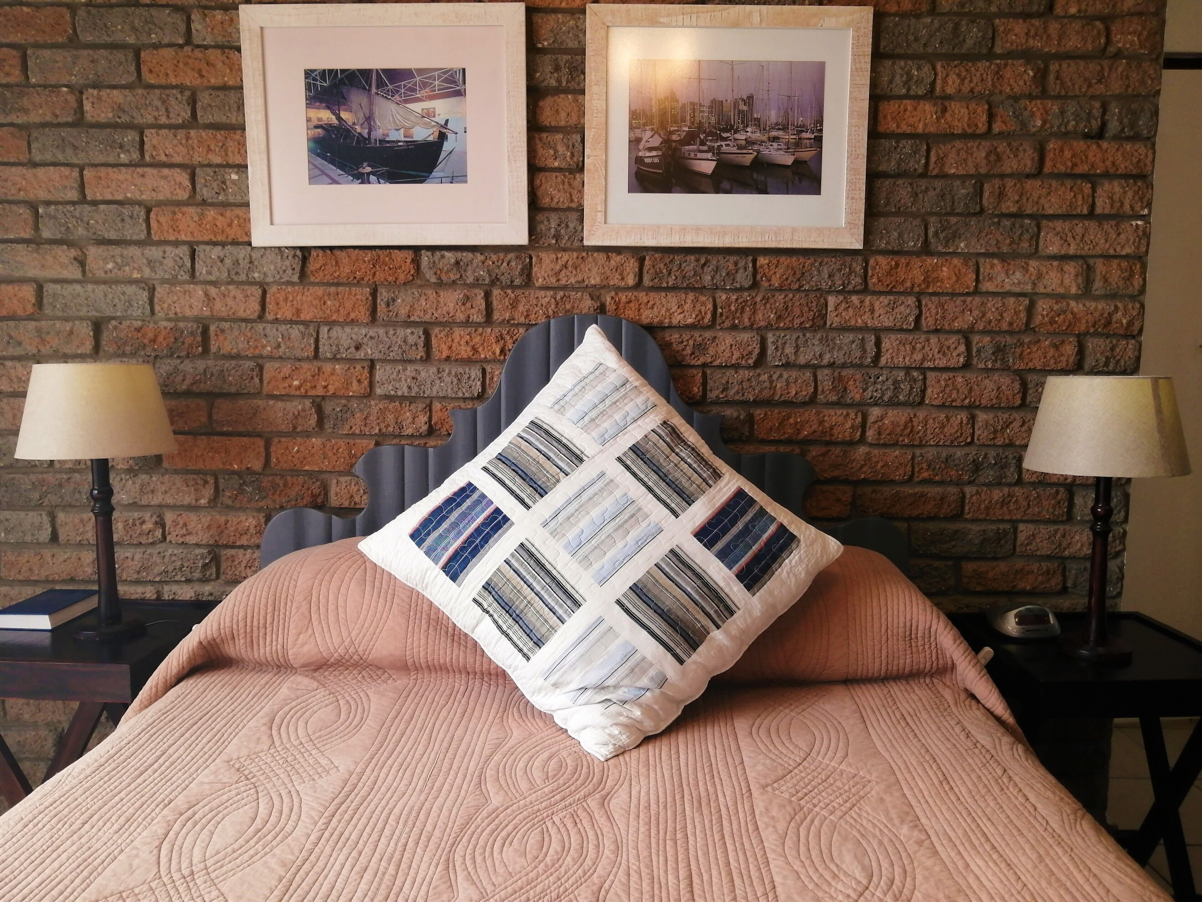 A hotel room with a brick wall, two framed pictures, a bed with a pink quilt, and two bedside lamps.