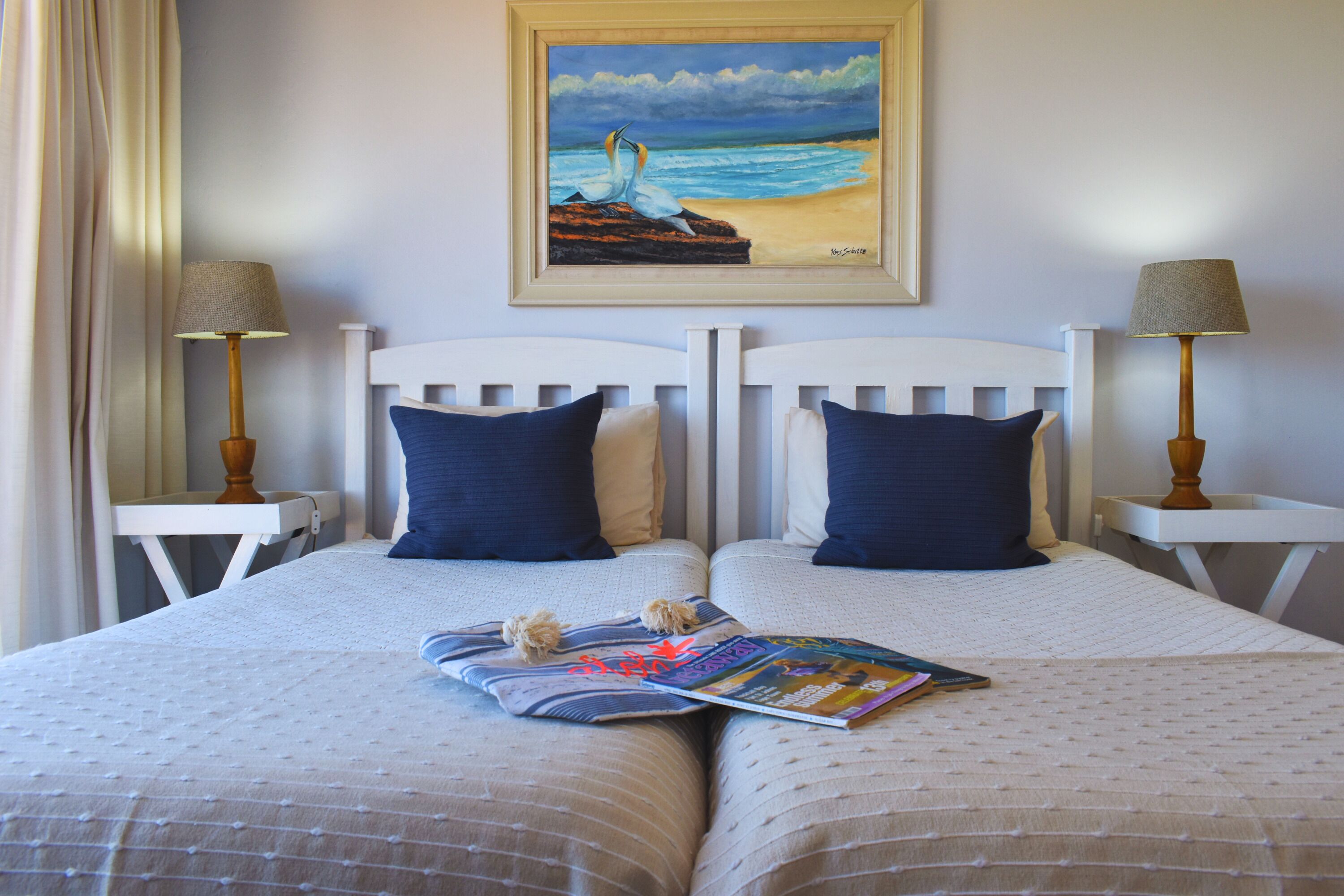 A double bed with a magazine and a small decorative item on it, flanked by two nightstands with lamps, a painting of a beach scene above, and a white headboard.