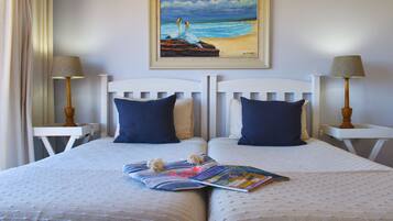 Una cama doble con una revista y un adorno decorativo, flanqueada por dos mesitas de noche con lámparas, un cuadro de una playa arriba y un cabecero blanco.