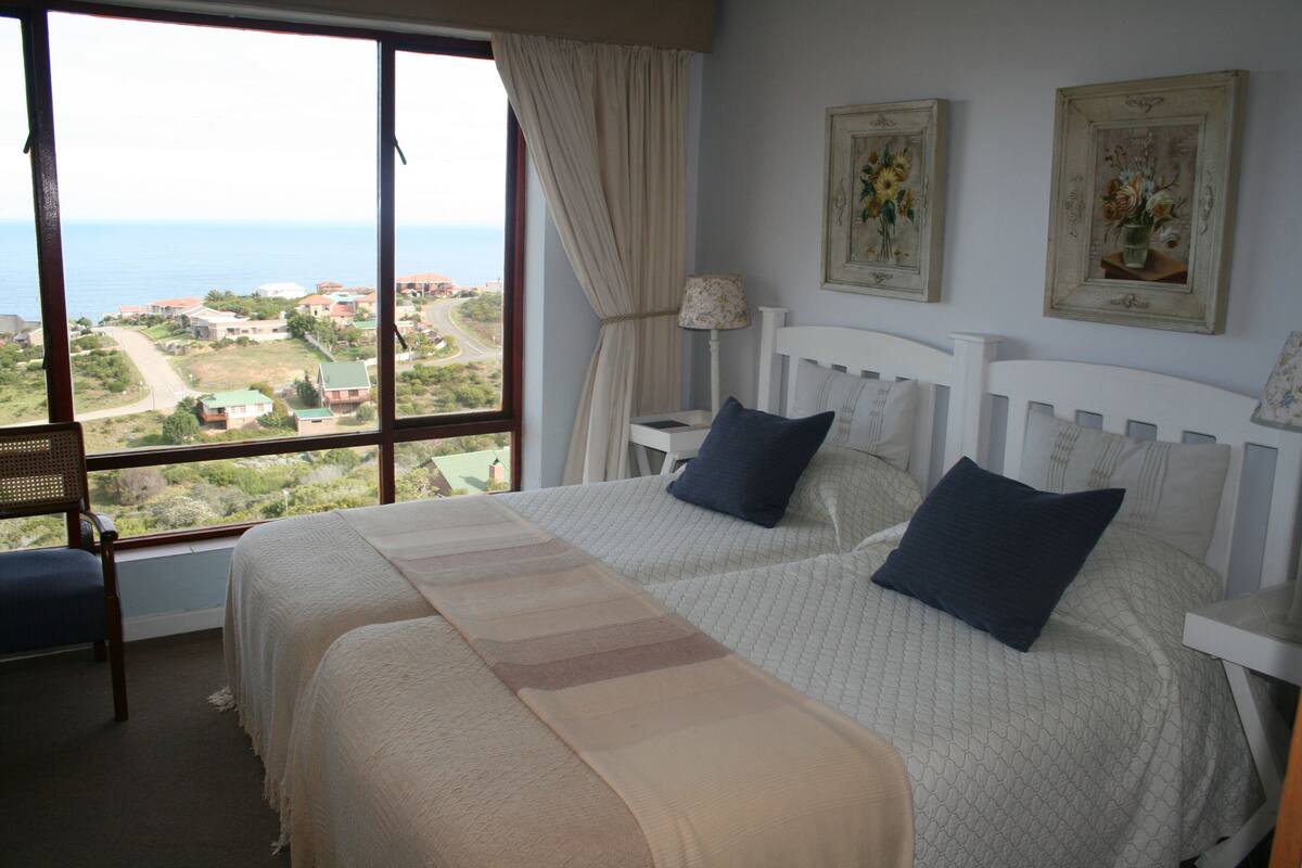 Un dormitorio con dos camas, un ventanal con vistas a la costa y dos cuadros en la pared.