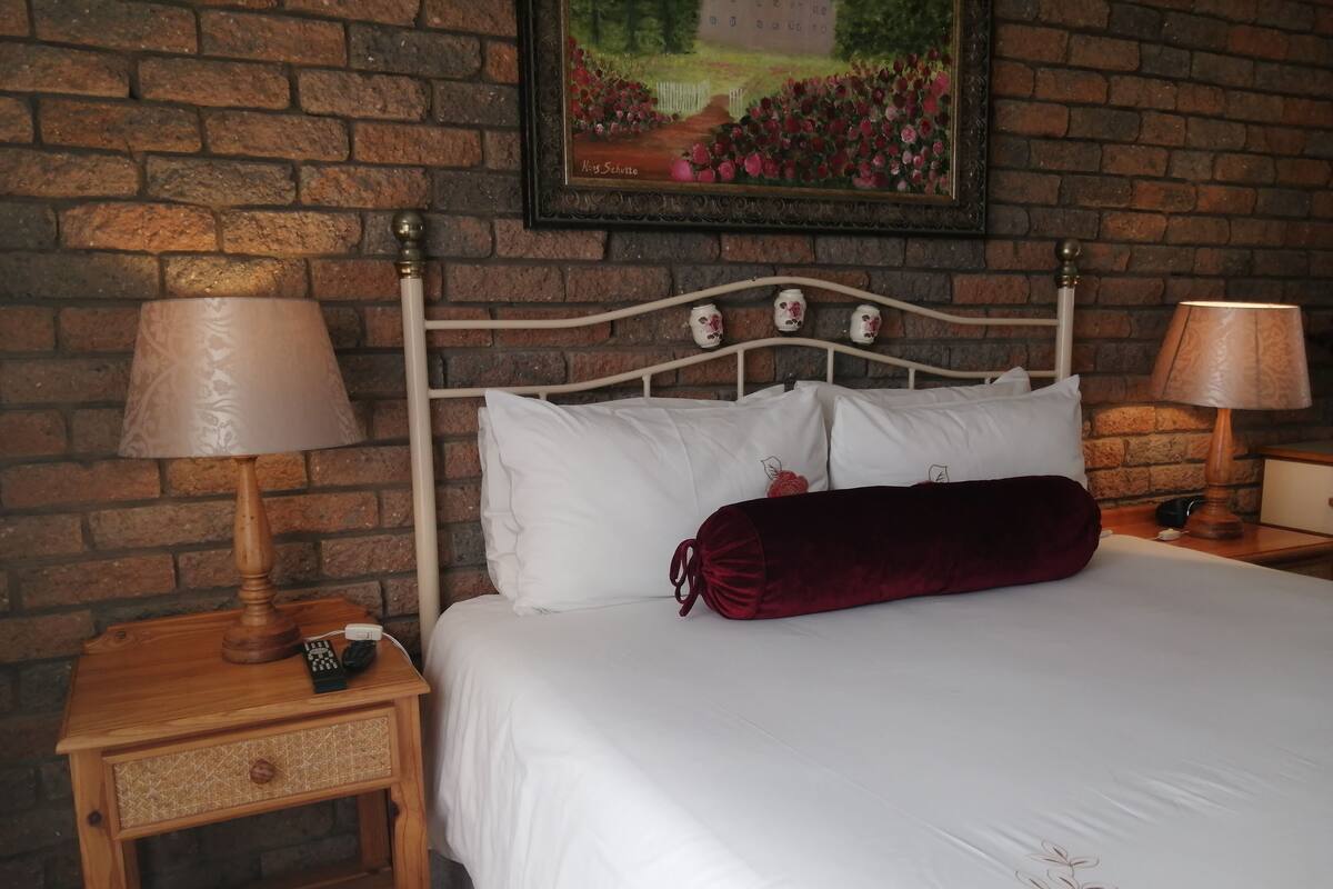 Una cama bien hecha con sábanas blancas y una almohada roja, una mesita de noche de madera con una lámpara, y una pared de ladrillo con un cuadro enmarcado.