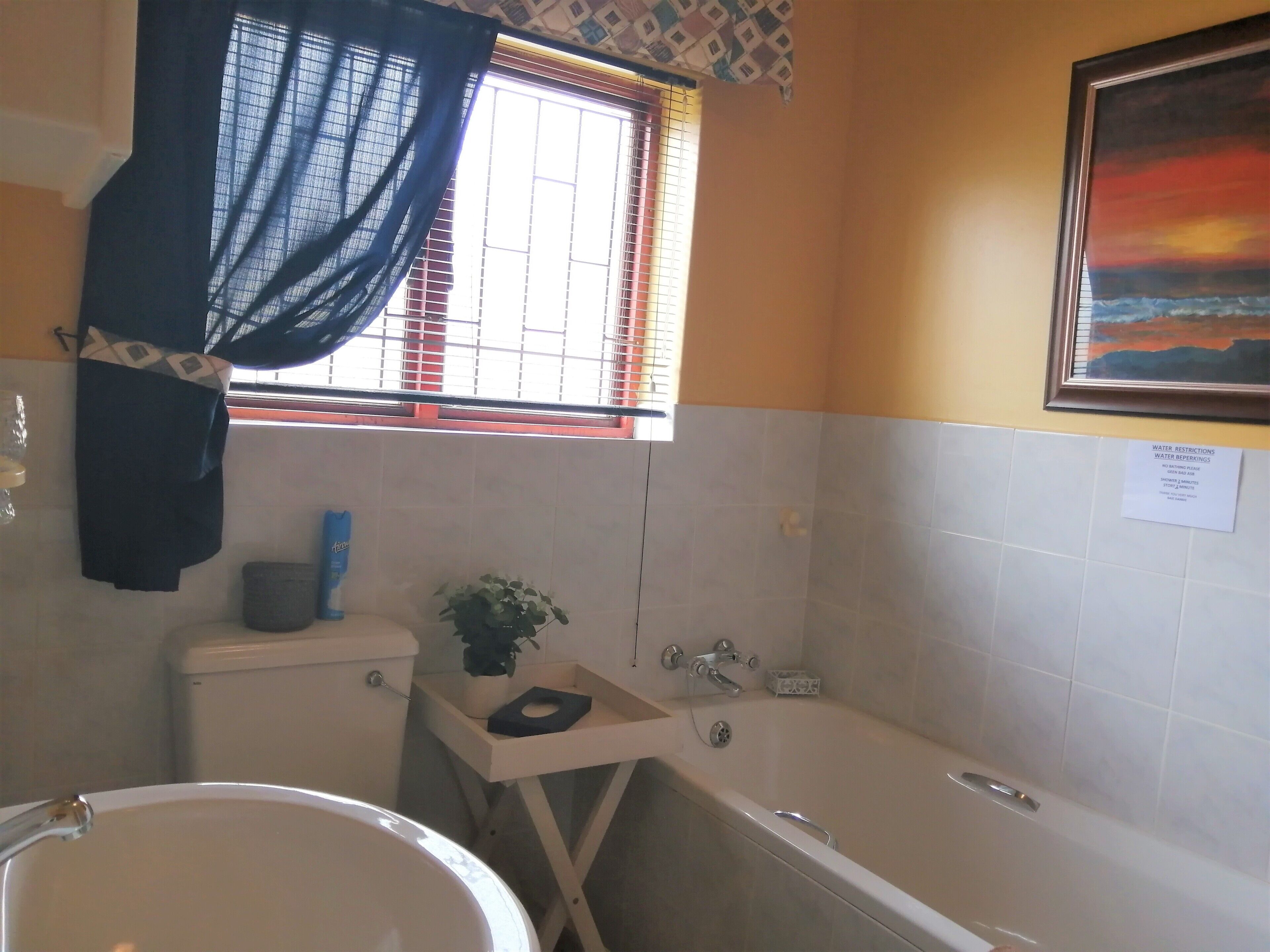 A bathroom with a bathtub, toilet, and sink. There is a window with a curtain, a plant on the side table, and a painting on the wall.