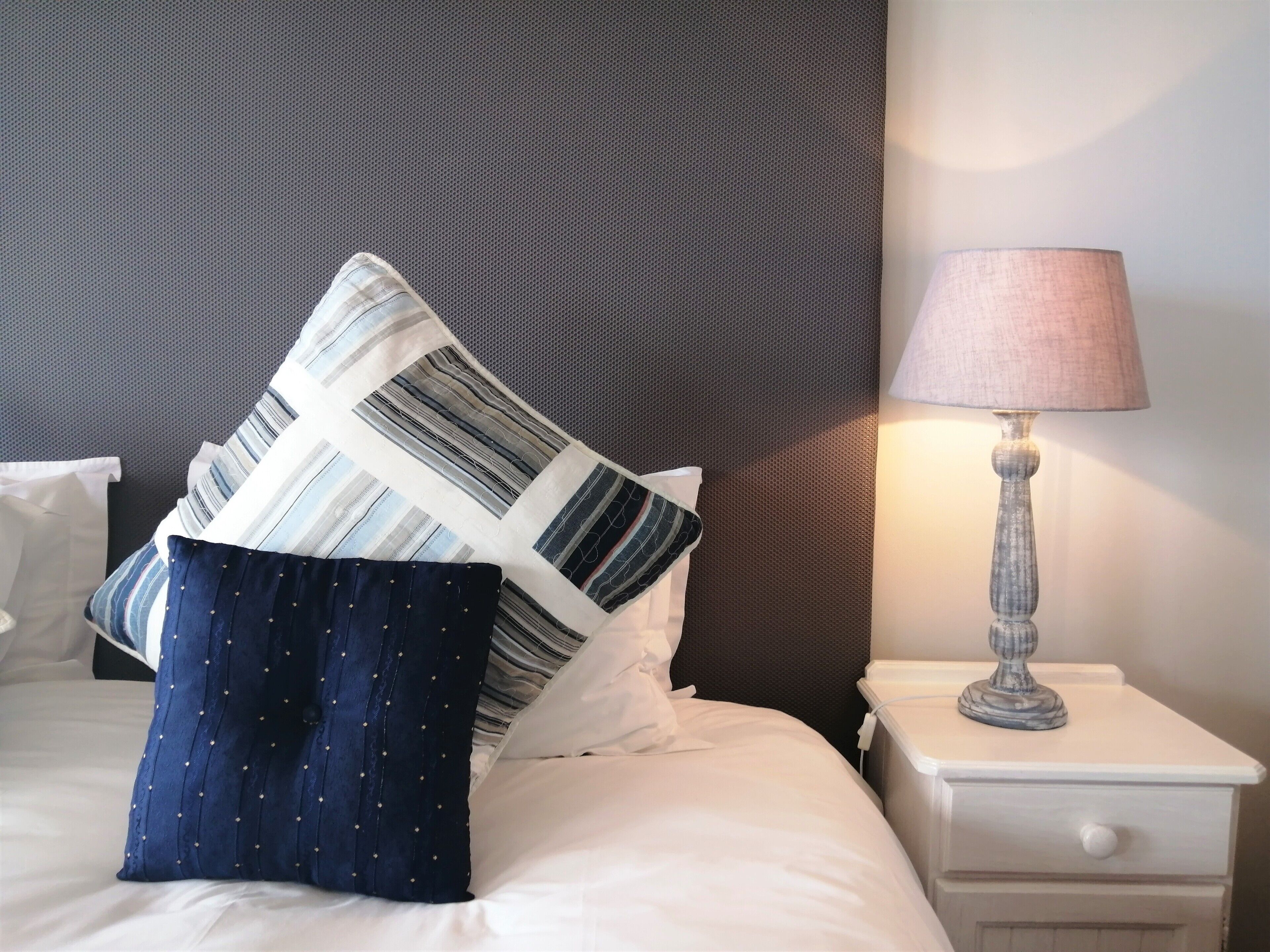 Un lit bien fait, agrémenté d’oreillers à rayures et à pois, une table de chevet blanche avec une lampe, et une tête de lit sombre à texture marquée.
