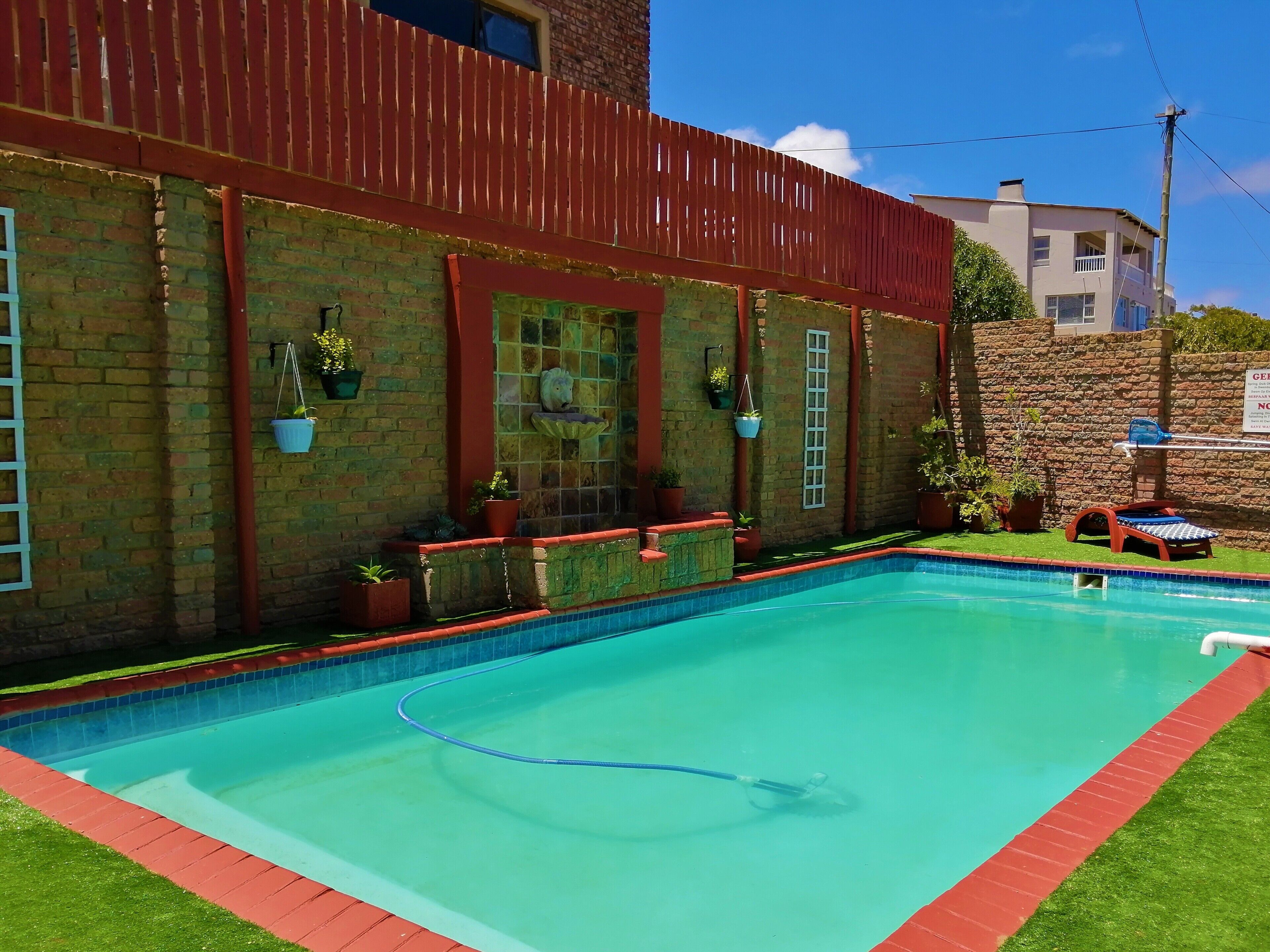 Una piscina con borde rojo, un muro de ladrillo con plantas colgantes y un muro de piedra al fondo.