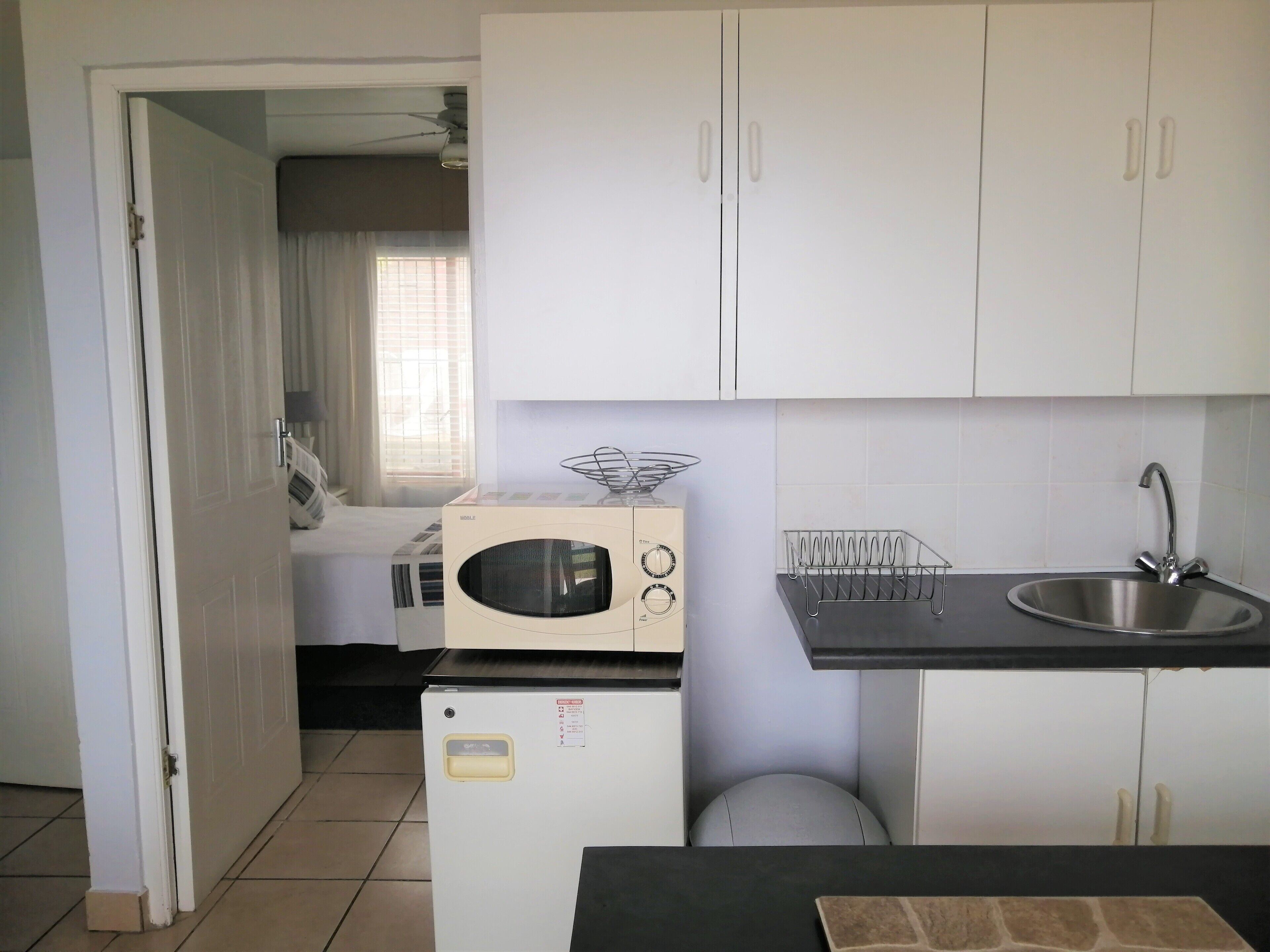 Een kleine keuken met witte kastjes, een magnetron, een kleine koelkast en een spoelbak.