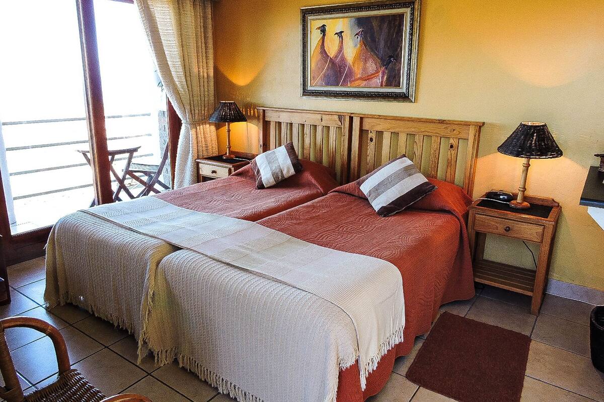 Una cama doble con dos almohadas, un cabecero de madera, una mesita de noche con lámpara, una silla y un cuadro en la pared.