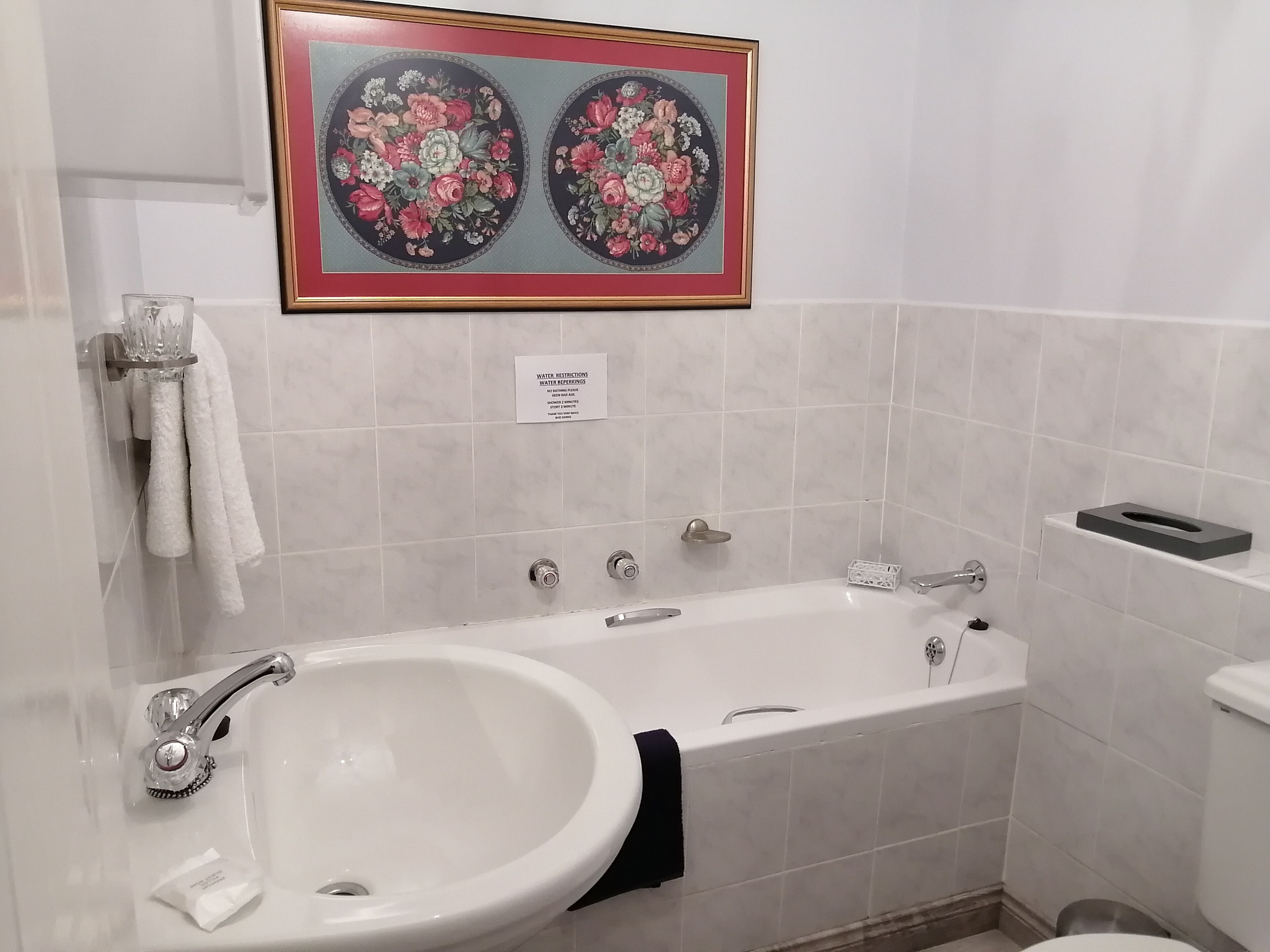 A bathroom with a sink, bathtub, and toilet. There is a framed floral artwork above the sink and a sign with water restrictions.