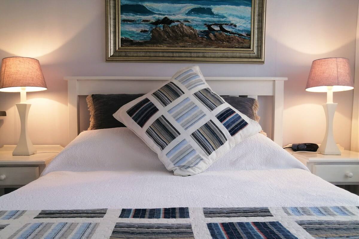 Una cama bien hecha con colcha, dos lámparas de noche, un cuadro enmarcado sobre la cama y una mesita de noche blanca con un teléfono.