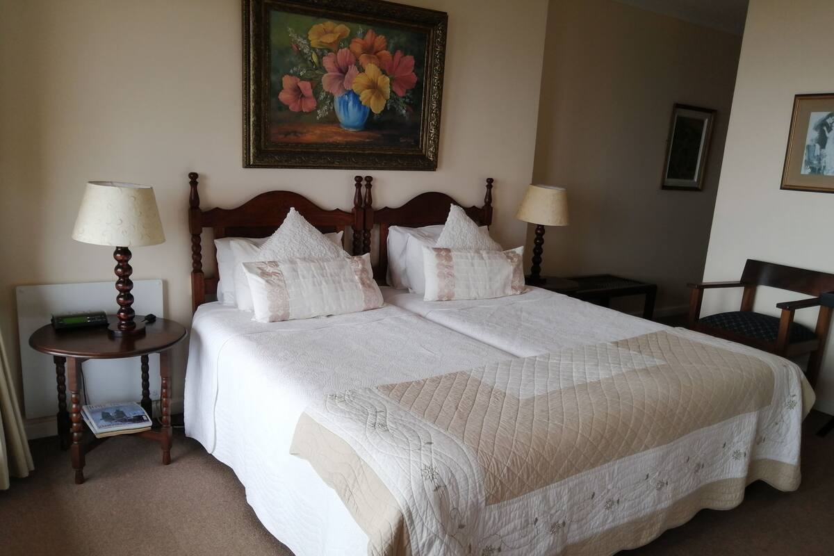 Una cama doble con colcha acolchada y almohadas, una mesita de noche con lámpara, un cuadro floral enmarcado en la pared y una silla en la esquina.