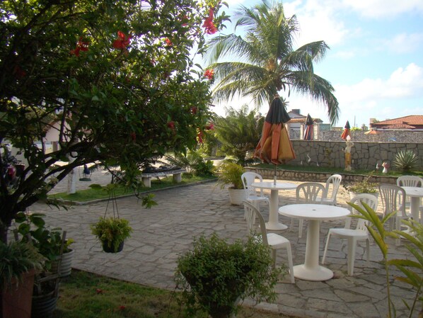 An outdoor patio with stone flooring, white plastic chairs, and tables. There are potted plants and a palm tree. - Beach Hotel Jacumã (Conde)