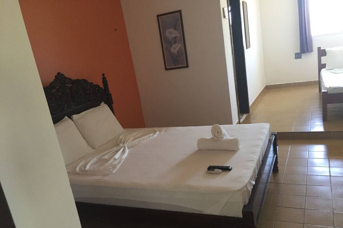 A neatly made bed with a remote control on it, a framed picture on the wall, and a chair in the background.