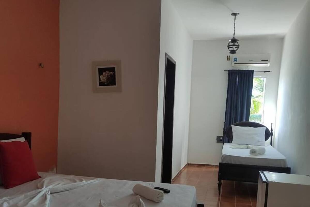 A hotel room with a large bed, a desk, and a chair. There is a window with curtains, a ceiling fan, and an air conditioning unit.
