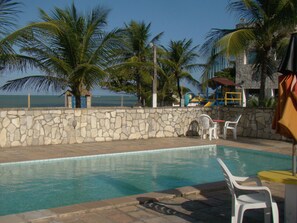 Un área de piscina con paredes de piedra, sillas de plástico blanco y palmeras.