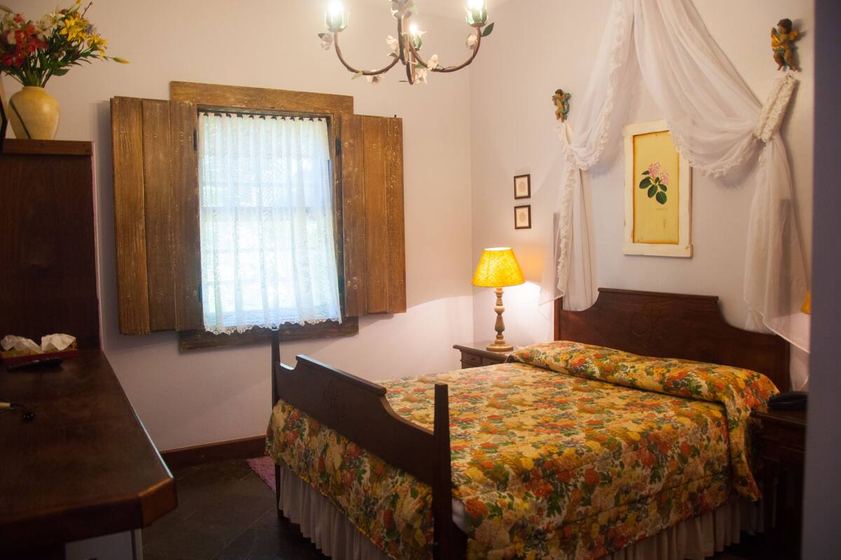 A bedroom with a wooden bed, a nightstand with a lamp, a window with curtains, and a wall-mounted picture.