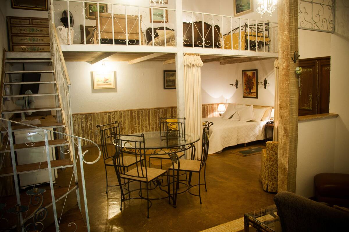 A room with a loft bed, a dining table with chairs, a staircase, and a small table with a chair.