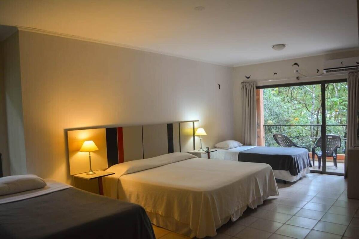 Camera d'albergo con due letti, un balcone con sedie e vista sul verde.