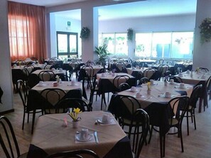 A spacious dining area with multiple tables and chairs, large windows with plants, and a door in the background. - Hotel Kennedy (Ravenna)