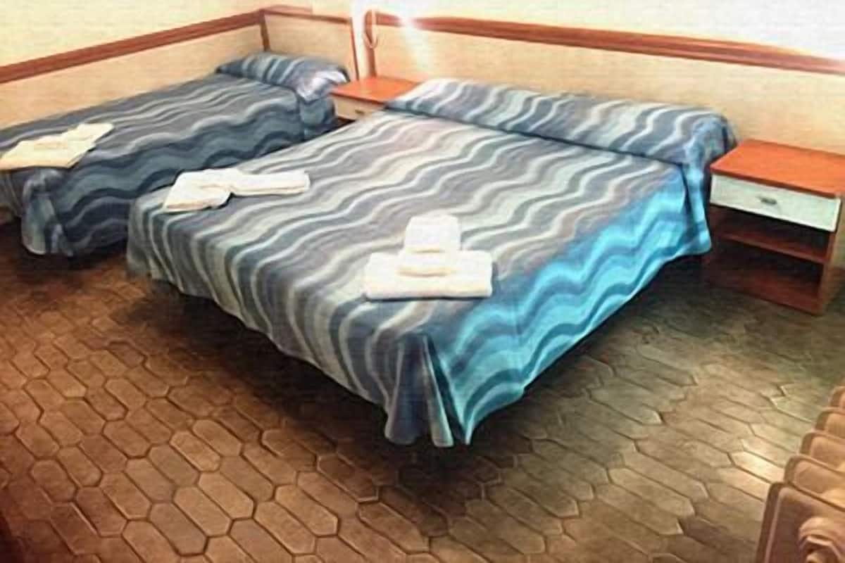 Habitación de hotel con dos camas, cada una con una colcha y cojines estampados, una mesita de noche con lámpara y suelo de madera.