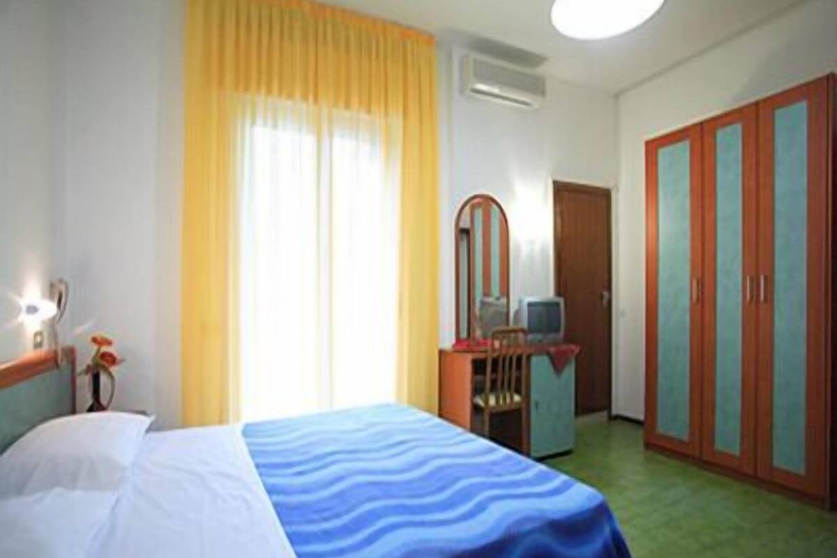 Habitación de hotel con cama, escritorio, silla, armario y ventana con cortinas.