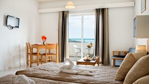 A hotel room with a bed, a desk, a chair, a table with a vase of flowers, and a view of the sea. - Sunny Hill Hotel Apartments (Chlorakas)