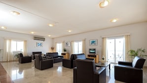A spacious living room with a large flat-screen TV, multiple seating areas, and a large window providing natural light. - Sunny Hill Hotel Apartments (Chlorakas)