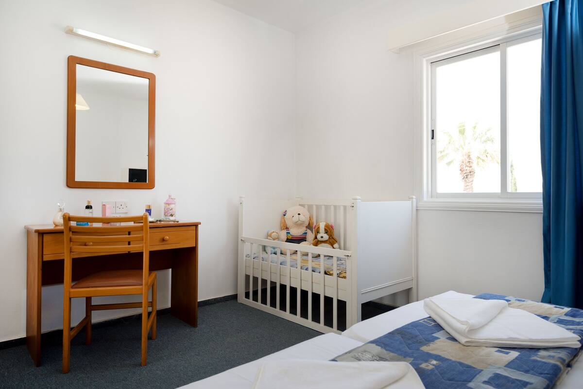 Ein Schlafzimmer mit einem Holztisch, einem Babybett mit Kuscheltieren, einem Bett mit einer blau-weißen Steudecke und einem Fenster mit blauen Vorhängen.