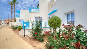 A white building with blue accents, a paved walkway, and various plants. - Sunny Hill Hotel Apartments (Chlorakas)