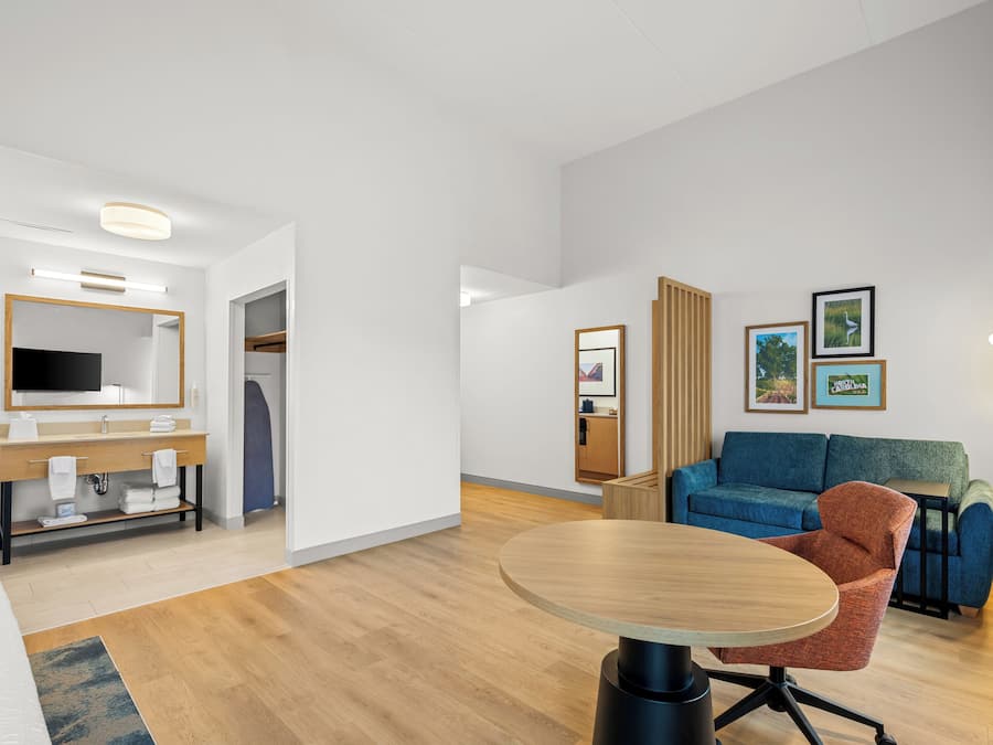 Habitación de hotel moderna con una mesa redonda de madera, un sofá, un televisor y un baño con espejo y lavamanos.