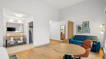 A modern hotel room with a round wooden table, a sofa, a television, and a bathroom with a mirror and sink.