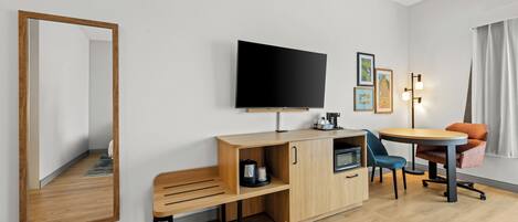 A modern hotel room with a flat-screen TV, a wooden bench, a small kitchenette, a round dining table with two chairs, and a large mirror.