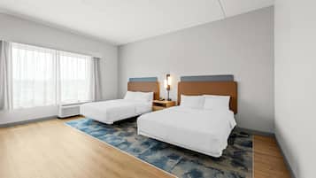 A hotel room with two beds, a large window with curtains, a standing heater, and a blue patterned rug.
