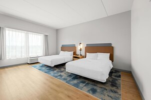 Habitación de hotel con dos camas, un ventanal grande con cortinas, un calentador portátil y una alfombra azul estampada.