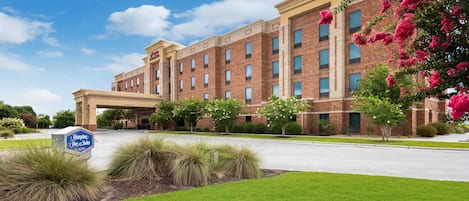 A large brick building with a clear sky, a sign for Hampton Inn & Suites, and well-maintained landscaping.