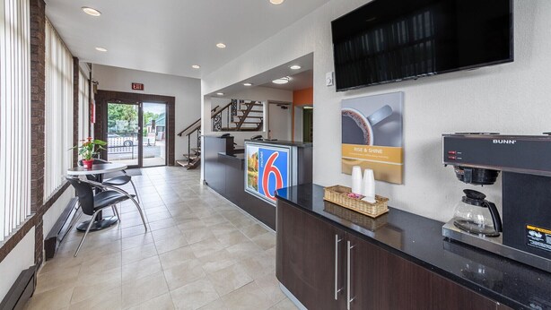 A hotel reception area with a coffee machine, a digital screen displaying room number 6, and a staircase leading to other floors. - Motel 6 Warminster, PA (Warminster)