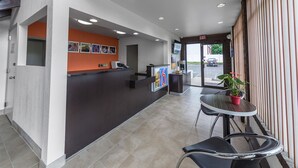 A hotel reception area with a dark reception desk, a small seating area with a table and chairs, and a view of the parking lot through the entrance. - Motel 6 Warminster, PA (Warminster)